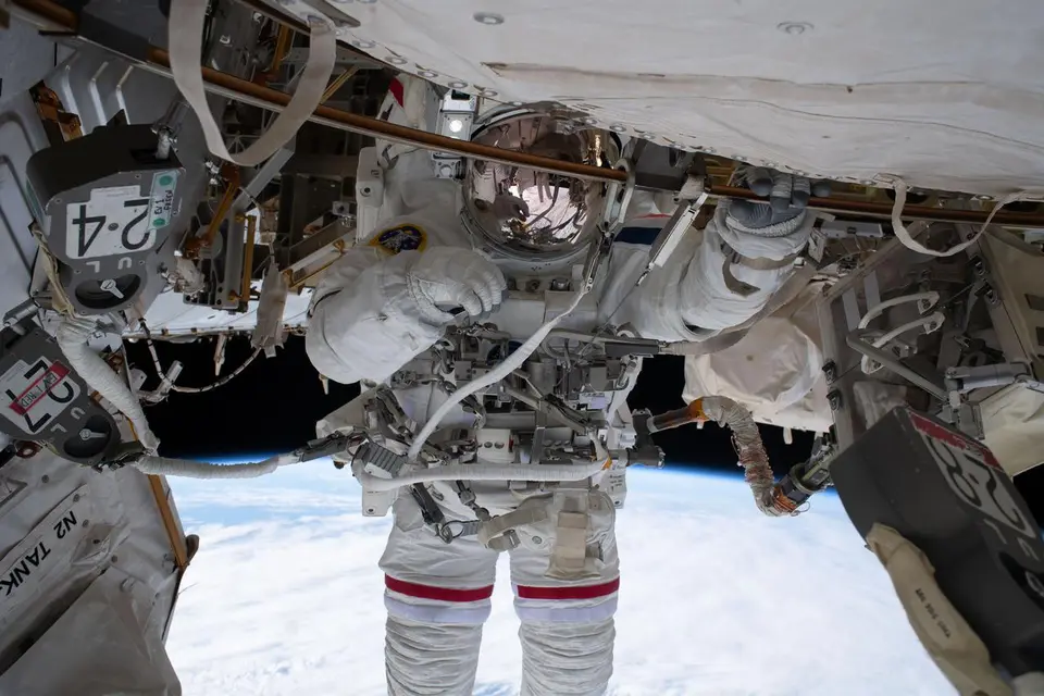 Astronauta em caminhada espacial preso por cabo à Estação Espacial, com a Terra iluminada embaixo
