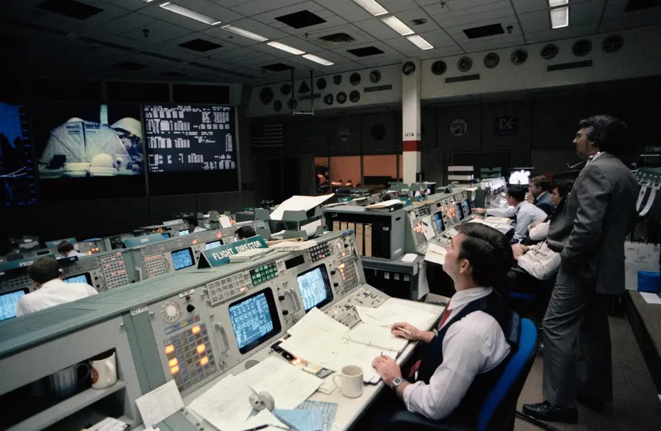 Sala de controle de missão com operadores diante de consoles e telas