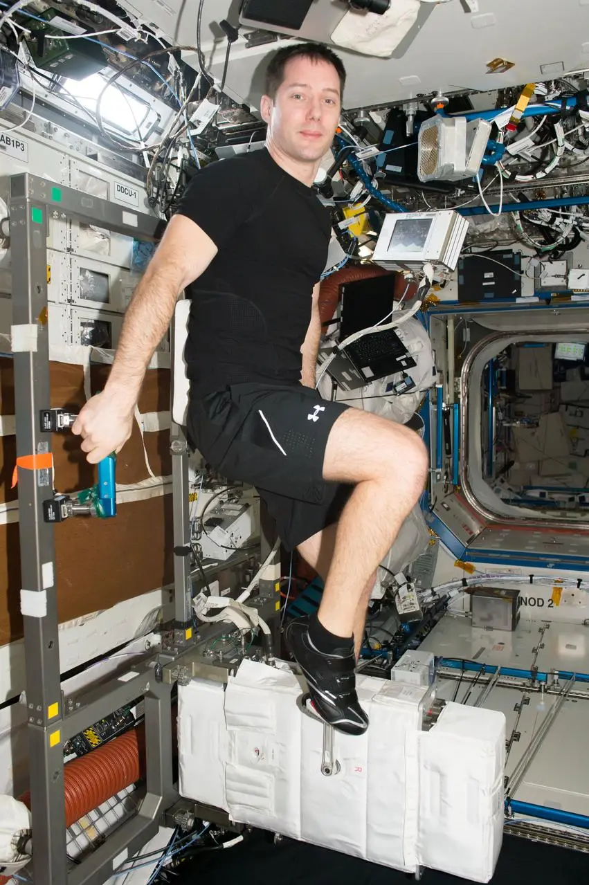 Astronauta pedalando em bicicleta ergométrica na Estação Espacial com equipamentos ao redor